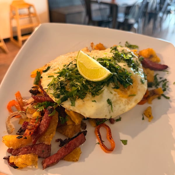 Breakfast plate with fried eggs, lime, cilantro, sausage, and peppers, in a restaurant setting.