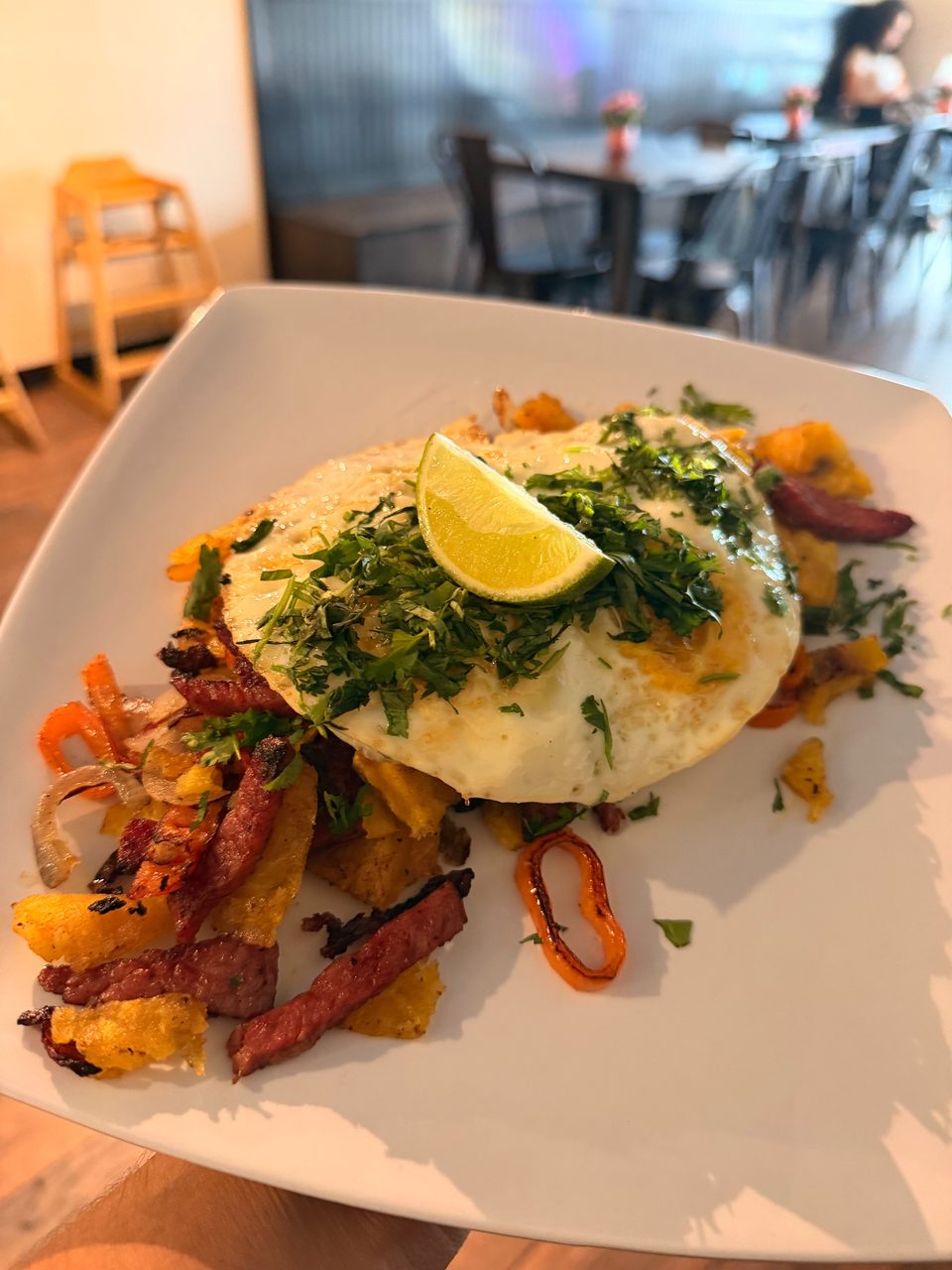 Breakfast plate with fried eggs, lime, cilantro, sausage, and peppers, in a restaurant setting.