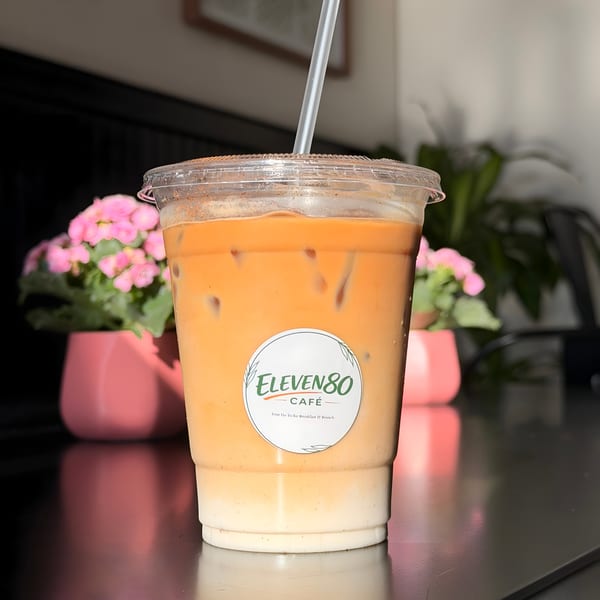 Iced coffee with milk and a straw, featuring the Eleven80 Cafe logo, on a dark table with pink flowers.
