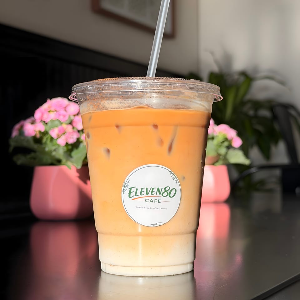 Iced coffee with milk and a straw, featuring the Eleven80 Cafe logo, on a dark table with pink flowers.