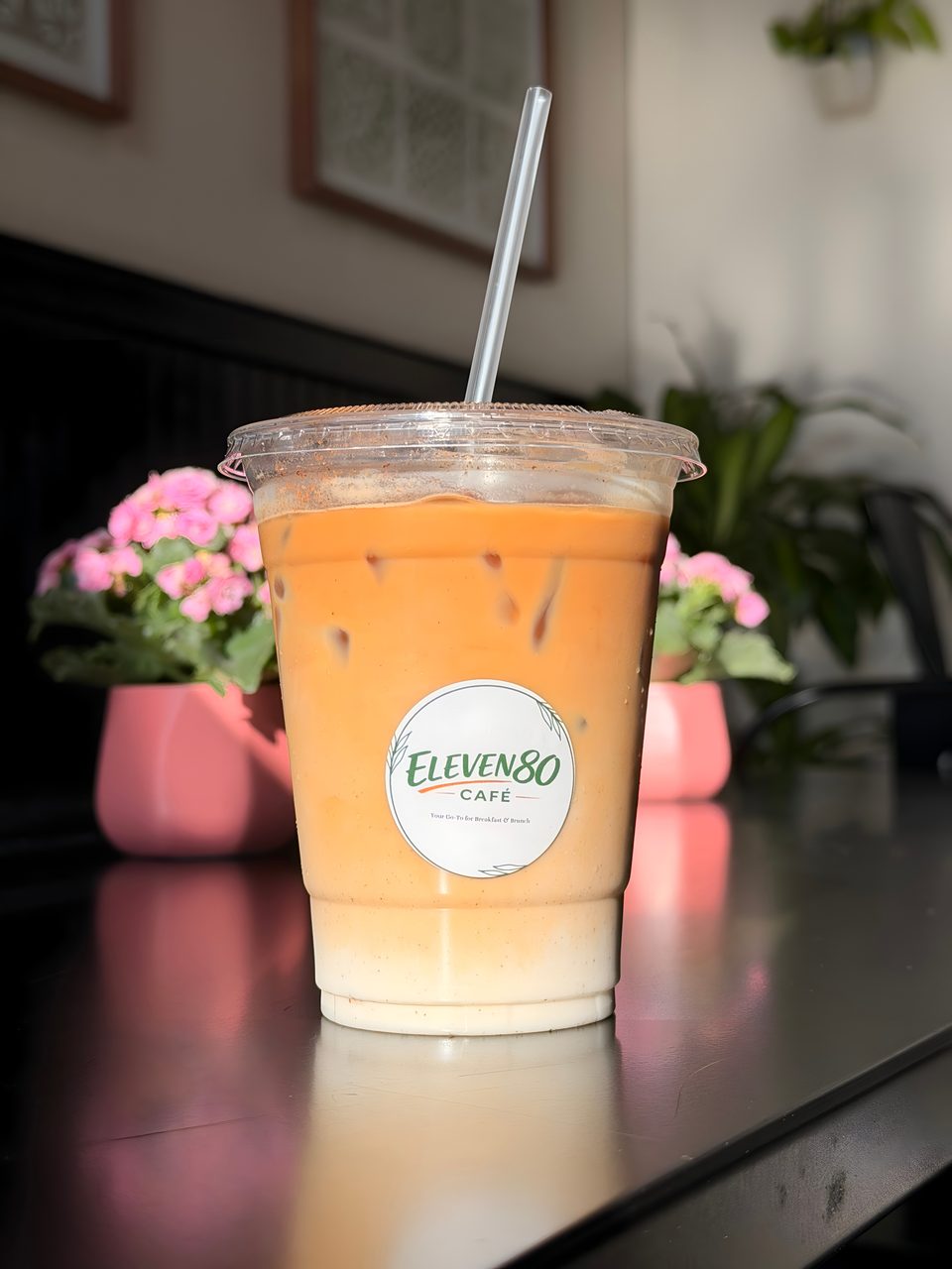 Iced coffee with milk and a straw, featuring the Eleven80 Cafe logo, on a dark table with pink flowers.